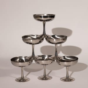 May include: A pyramid of seven stainless steel champagne coupe glasses. Each glass has a shallow, rounded bowl and a slender stem. The glasses are arranged in a stacked formation, creating a visually appealing display.