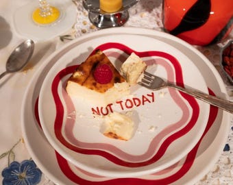 May include: A plate with a slice of cheesecake topped with a raspberry and the words "NOT TODAY" in red. The plate has a red and pink border and sits on a white plate. A fork rests on the plate.