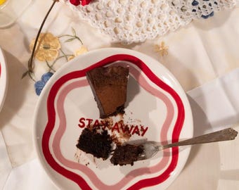 May include: A white plate with a red and pink wavy border, featuring a slice of chocolate cake and the words "STAY AWAY". A fork rests on the plate, with a piece of cake missing. The scene includes a bowl of berries and a red flower.