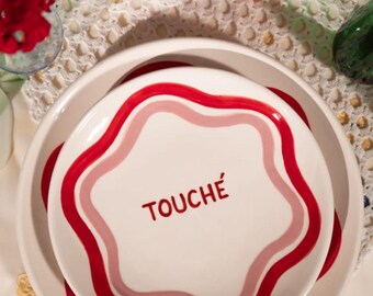 May include: White ceramic plates with a red and pink wavy border. The top plate has the word "TOUCHÉ" in red. The plates are stacked on a white lace tablecloth, with other serving dishes and flowers in the background.