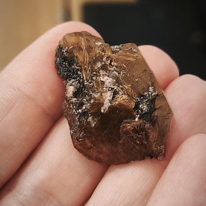 Könnte beinhalten: Ein rohes, bernsteinfarbenes Mineralexemplar mit dunklen Einschlüssen und weißen kristallinen Flecken. Der Stein wird in einer Hand gehalten, wodurch seine natürliche Textur und Farbvariationen hervorgehoben werden. Das Mineral scheint ein roher, unpolierter Edelstein zu sein.