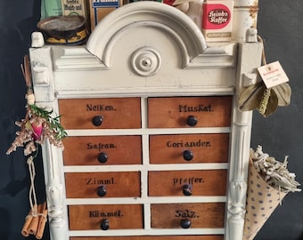 May include: Antique spice rack with twelve drawers labeled with spice names in German. The white-painted wooden cabinet is topped with vintage spice containers and small bags. Dried flowers and cinnamon sticks add to the rustic charm.