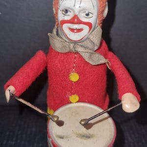 Puede incluir: Payaso de juguete vintage de cuerda, con chaqueta de fieltro roja y botones amarillos, tocando un tambor. El payaso tiene una cara pintada con una sonrisa roja, un sombrero negro y pelo naranja. El tambor es beige con detalles rojos y blancos.