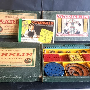 Puede incluir: Juego de construcción vintage Marklin Metallbaukasten con varias cajas verdes y doradas. Las cajas presentan el logotipo de Marklin. El juego incluye varias piezas metálicas, engranajes y folletos de instrucciones. La caja abierta revela una variedad de componentes coloridos.