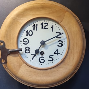 Puede incluir: Un reloj redondo de madera clara con esfera blanca y números negros. El reloj tiene manecillas negras y una llave de metal insertada en el lado izquierdo. El marco del reloj es de color marrón claro.