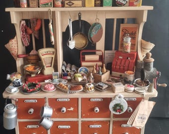 May include: A miniature kitchen display with a wooden cabinet featuring orange drawers and a shelf filled with tiny food items, vintage packaging, and kitchen tools. Includes a scale, cash register, and hanging pots.
