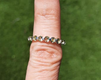Op de afbeelding: Een zilveren ring met een rij kleine, ronde, lichtgroene edelstenen. De ring wordt op een vinger gedragen, met een groene achtergrond zichtbaar. De ring is een sieraad.