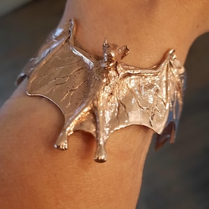 May include: A gold-toned metal bracelet shaped like a bat with its wings spread wide. The bat is detailed with textured wings and a realistic body.