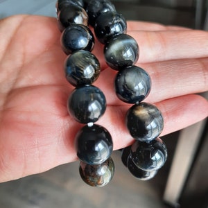 Puede incluir: Dos pulseras de cuentas negras y doradas con un patrón de ojo de tigre. Las pulseras están hechas de cuentas redondas y están apiladas una encima de la otra.