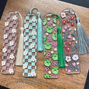 May include: Four clear acrylic bookmarks with different designs and tassels. The bookmarks feature a checkered pattern with white flowers, a frog pattern with pink bows, a floral pattern with red flowers, and a checkered pattern with white flowers.