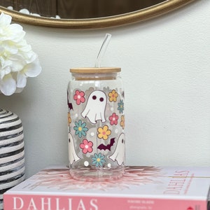 May include: A clear glass reusable water bottle with a bamboo lid and a straw. The bottle has a design of white ghosts, bats, and colorful flowers. The bottle is sitting on top of two books.