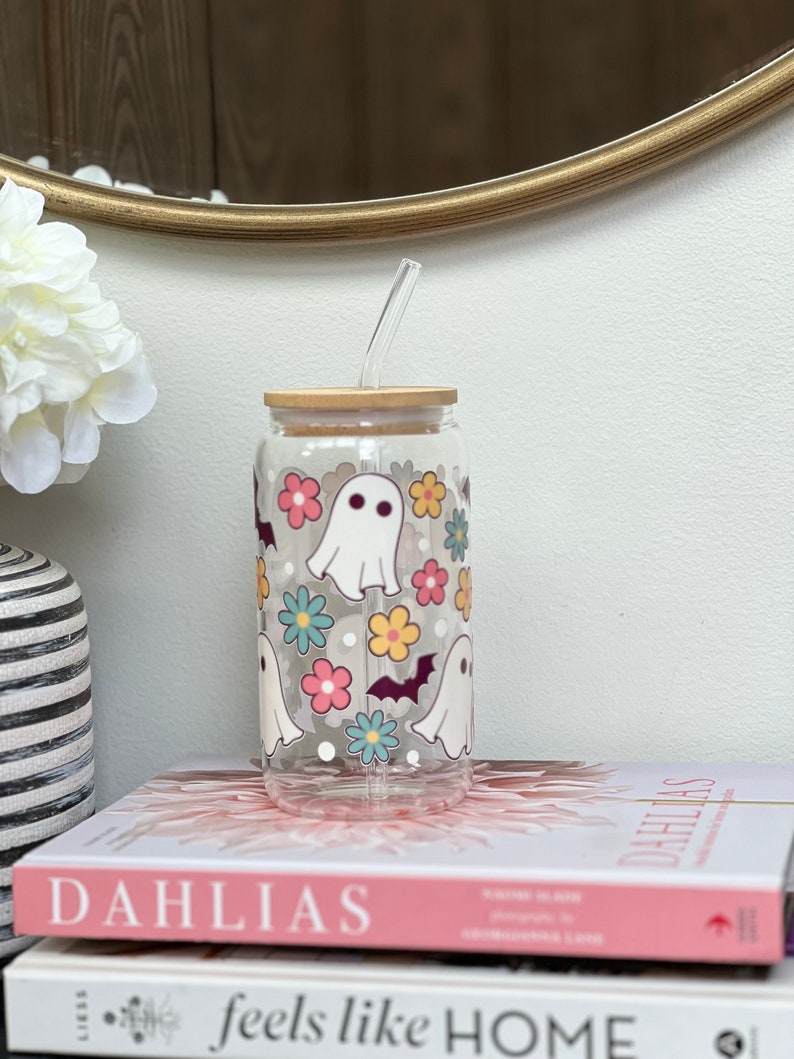 May include: A clear glass reusable water bottle with a bamboo lid and a straw. The bottle has a design of white ghosts, bats, and colorful flowers. The bottle is sitting on top of two books.