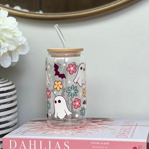 May include: A clear glass reusable cup with a bamboo lid and a straw. The cup has a Halloween theme with ghosts and bats printed on it. The cup is sitting on top of two books.