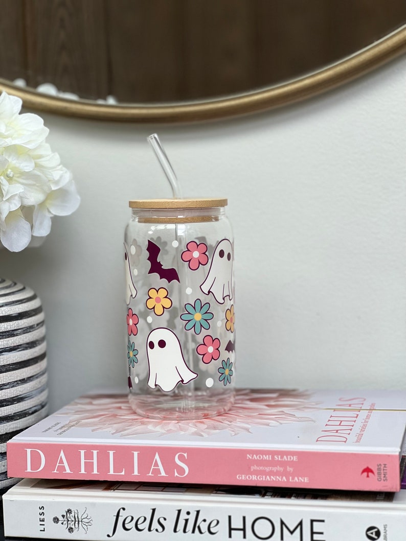 May include: A clear glass reusable cup with a bamboo lid and a straw. The cup has a Halloween theme with ghosts and bats printed on it. The cup is sitting on top of two books.