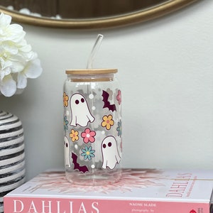 May include: A clear glass reusable water bottle with a wooden lid and a straw. The bottle has a whimsical design featuring ghosts, bats, and flowers in pink, yellow, blue, and purple.