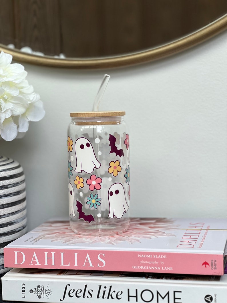 May include: A clear glass reusable water bottle with a wooden lid and a straw. The bottle has a whimsical design featuring ghosts, bats, and flowers in pink, yellow, blue, and purple.