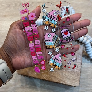 Hand holding three acrylic bookmarks with tassels: pink “Hot Girls Read Books” with hearts and flowers, pastel “Book Girl Era” with stacked books and daisies, and red “Smutty Book Club” with chili peppers, flames, and handcuffs. Fun, aesthetic reading accessories for book lovers, coquette readers, and book clubs.