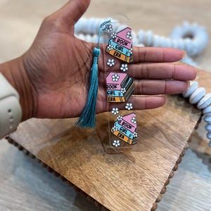 Hand holding pastel acrylic bookmark with teal tassel featuring stacked books labeled “Book Girl Era” with white daisies. Aesthetic acrylic bookmark for cozy readers, book clubs, and unique bookish gifts.