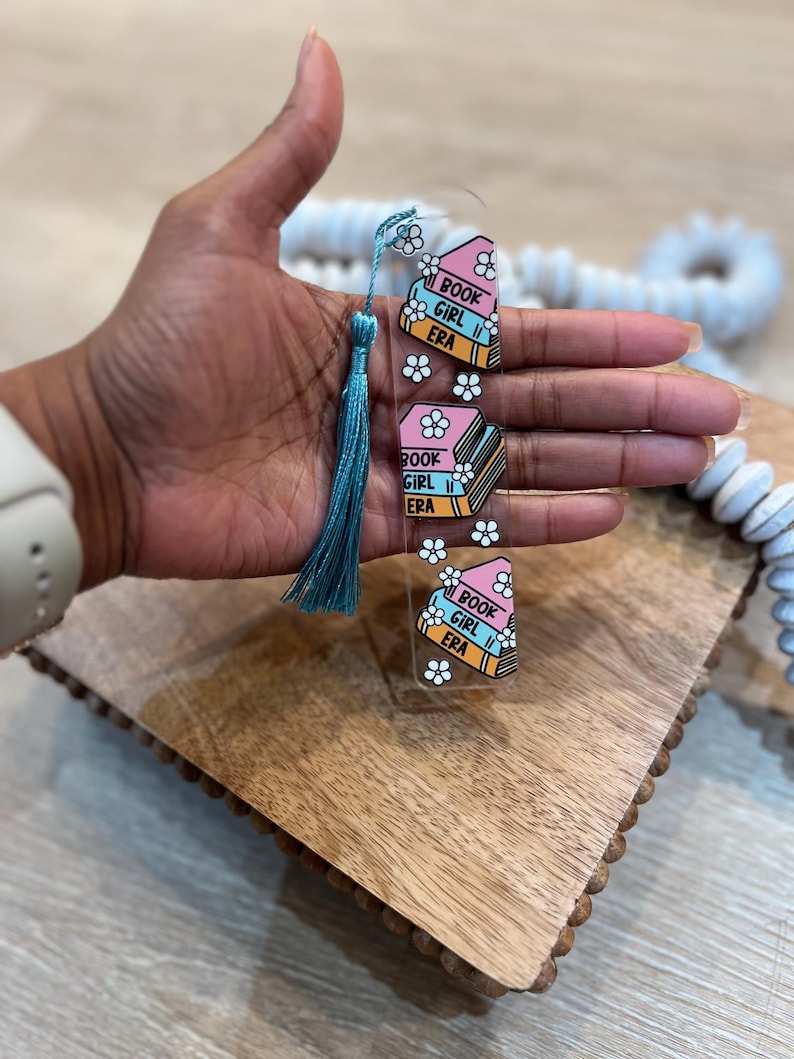 Hand holding pastel acrylic bookmark with teal tassel featuring stacked books labeled “Book Girl Era” with white daisies. Aesthetic acrylic bookmark for cozy readers, book clubs, and unique bookish gifts.