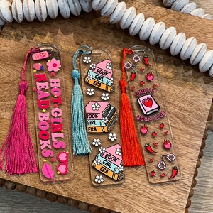 Flat lay of three acrylic bookmarks with tassels: pink “Hot Girls Read Books,” pastel “Book Girl Era” with daisies, and red “Smutty Book Club” with spicy motifs. Perfect gifts for book lovers, book clubs, romance readers, and coquette aesthetic stationery fans.