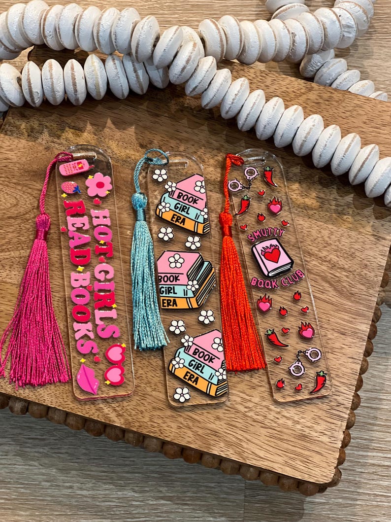Flat lay of three acrylic bookmarks with tassels: pink “Hot Girls Read Books,” pastel “Book Girl Era” with daisies, and red “Smutty Book Club” with spicy motifs. Perfect gifts for book lovers, book clubs, romance readers, and coquette aesthetic stationery fans.