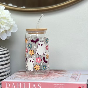 May include: A clear glass tumbler with a wooden lid and a straw. The tumbler has a Halloween-themed design with ghosts, bats, and flowers in pink, yellow, blue, and green.