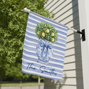 Puede incluir: Una bandera de jardín a rayas azules y blancas con una ilustración de acuarela de un jarrón azul y blanco con un limonero en su interior. El texto "The Smiths" está impreso en la parte inferior de la bandera.