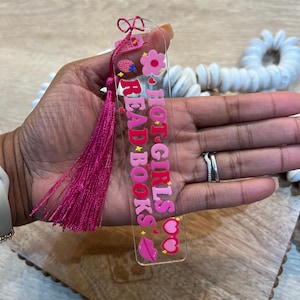 Close-up of hand holding pink acrylic bookmark with tassel that says “Hot Girls Read Books,” decorated with hearts, lips, flowers, and retro cellphone. Cute and trendy reading accessory for coquette book girls, romance readers, and book club gifts.