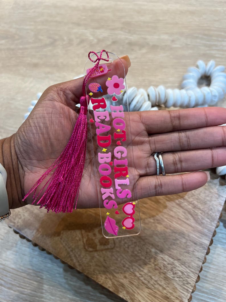 Close-up of hand holding pink acrylic bookmark with tassel that says “Hot Girls Read Books,” decorated with hearts, lips, flowers, and retro cellphone. Cute and trendy reading accessory for coquette book girls, romance readers, and book club gifts.