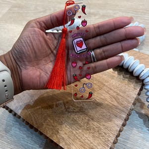 Hand holding red acrylic bookmark with tassel that says “Smutty Book Club,” decorated with chili peppers, flames, hearts, and handcuffs. Playful bookish gift for romance readers, spice lovers, and book club members.