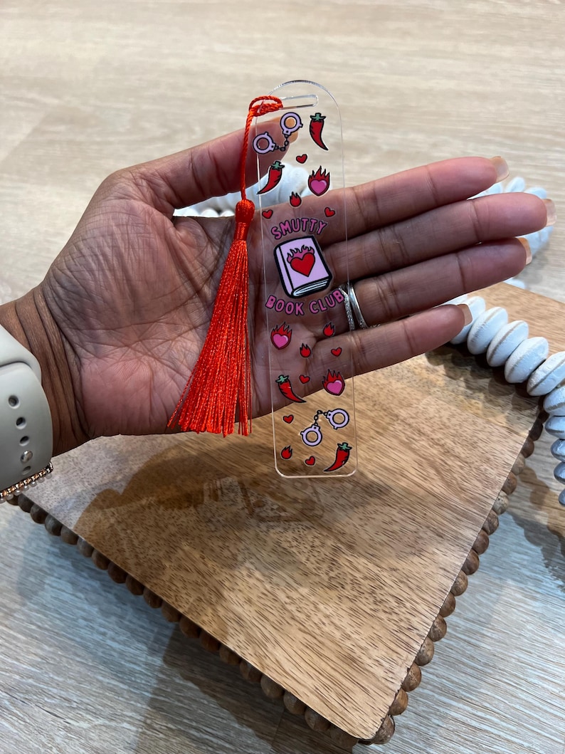 Hand holding red acrylic bookmark with tassel that says “Smutty Book Club,” decorated with chili peppers, flames, hearts, and handcuffs. Playful bookish gift for romance readers, spice lovers, and book club members.