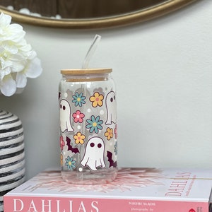 May include: A clear glass reusable water bottle with a bamboo lid and a straw. The bottle has a whimsical design featuring white ghosts, bats, and colorful flowers.