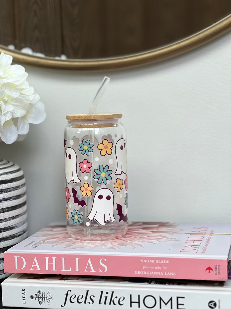 May include: A clear glass reusable water bottle with a bamboo lid and a straw. The bottle has a whimsical design featuring white ghosts, bats, and colorful flowers.