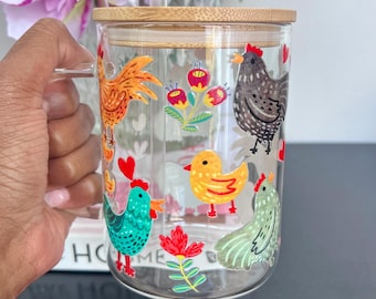 Pode incluir: Uma caneca de vidro transparente com uma alça, com ilustrações coloridas de galinhas, galos e flores. A caneca tem uma tampa de bambu e um canudo de vidro. A palavra "HOME" é parcialmente visível em um livro atrás da caneca.