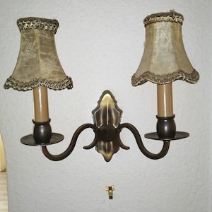 Könnte beinhalten: Eine Wandleuchte mit zwei Armen und zwei beigefarbenen Lampenschirmen mit goldenen Verzierungen. Die Leuchte hat eine dekorative Bronze-Oberfläche und ein geschwungenes Design.
