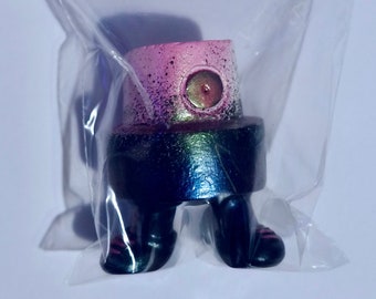 May include: A limited edition art toy sculpture of a spray can cap with legs, packaged in a clear plastic bag. The cap is pink and black with a metallic finish. The packaging includes the text "Street Toys" and "Mr. Faiga."