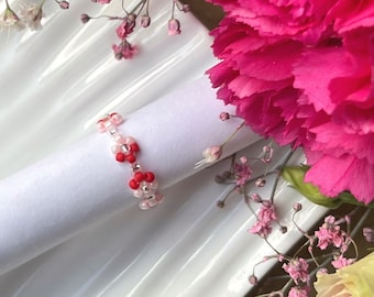 Può includere: Un anello per tovaglioli bianco con perline rosa e rosse. L'anello è su un piatto bianco con fiori rosa.