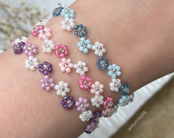 Può includere: Diversi braccialetti di fiori di perline nei toni del viola, rosa, bianco e blu. I braccialetti sono indossati al polso, mettendo in risalto il delicato design floreale. Le piccole perline creano un accessorio affascinante e femminile.
