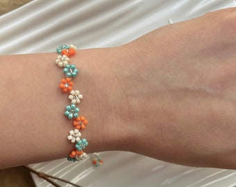 Può includere: Un braccialetto con piccole perline a forma di fiore di colore arancione, bianco e turchese.
