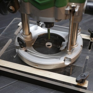 Puede incluir: Primer plano de una fresa de router fijada a un router, montada en una plantilla de acrílico transparente con una base de madera. La fresa de router está centrada en la plantilla y está lista para ser utilizada en proyectos de carpintería.