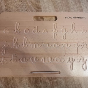 Può includere: Tavola di tracciamento dell'alfabeto in legno con uno stilo per esercitarsi nella scrittura. La tavola presenta le lettere dell'alfabeto in corsivo, con un solco per tracciare ogni lettera. La tavola è etichettata "Mimi Montessori".