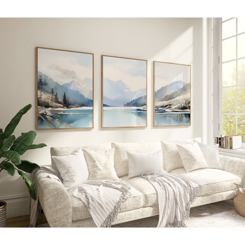 May include: Three framed watercolour paintings depicting a mountain lake scene with blue and grey tones. The paintings are hung on a white wall above a white sofa with throw pillows and a blanket.