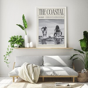 May include: A framed monochrome print titled "THE COASTAL" showing two people on surfboards in the sea. The print is displayed above a light grey sofa with a wooden frame, with plants and decorative items.