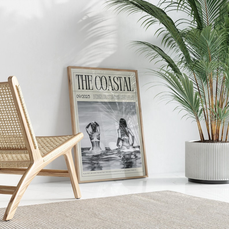 May include: A framed print titled "THE COASTAL" with a black and white image of two people in the sea. The print is in a light wood frame and is leaning against a white wall. A wooden chair and a potted plant are also in the scene.