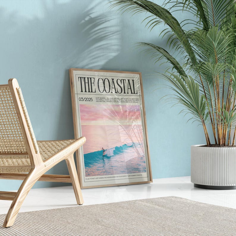 May include: A framed poster titled "THE COASTAL" with a beach scene, a surfer on a wave, and the date 03/2025. The poster is leaning against a light blue wall next to a wooden chair and a potted plant.