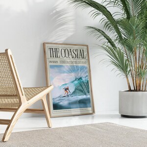 May include: A framed poster titled "THE COASTAL" featuring a surfer riding a wave. The poster has a light wood frame and is leaning against a white wall. A wooden chair and a potted plant are in the foreground.