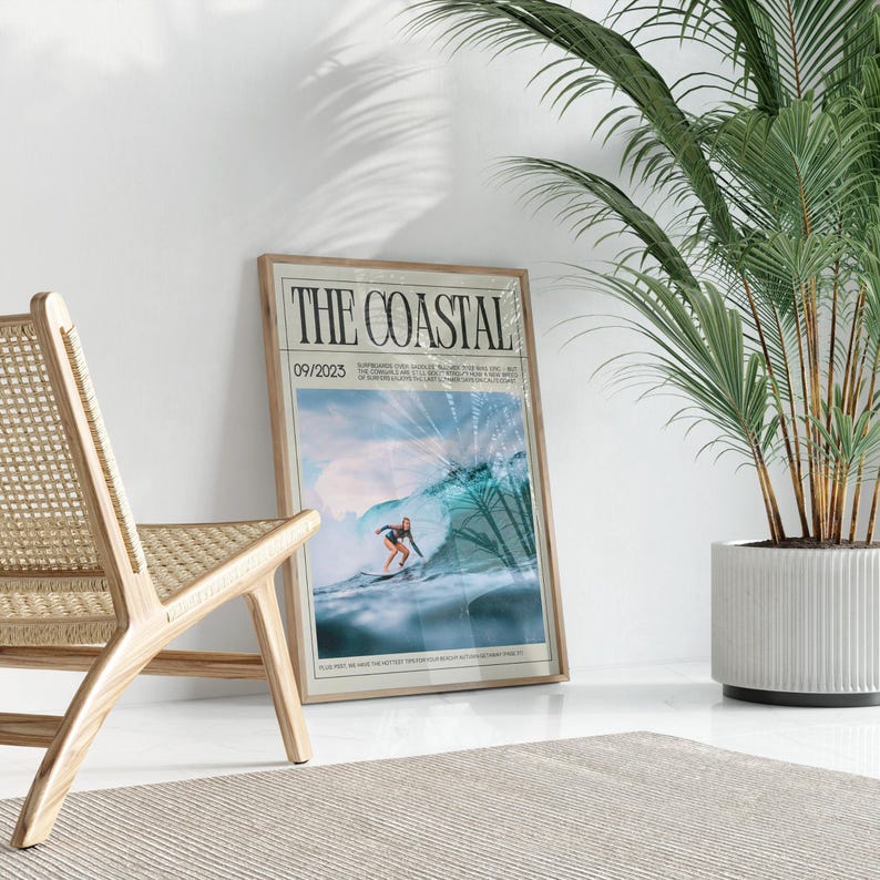 May include: A framed poster titled "THE COASTAL" featuring a surfer riding a wave. The poster has a light wood frame and is leaning against a white wall. A wooden chair and a potted plant are in the foreground.