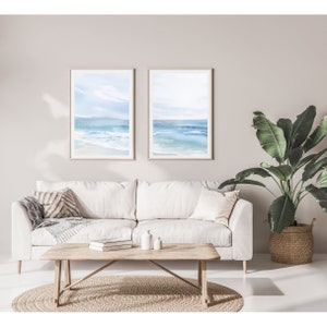 May include: Two framed watercolor paintings of a seascape with blue water and white sand. The paintings are hanging on a light beige wall above a white sofa with pillows and a wooden coffee table.