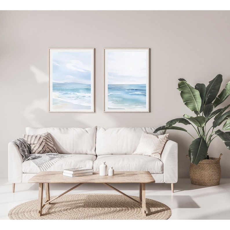 May include: Two framed watercolor paintings of a seascape with blue water and white sand. The paintings are hanging on a light beige wall above a white sofa with pillows and a wooden coffee table.