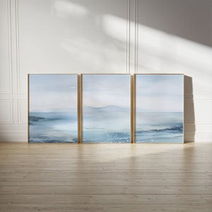 Puede incluir: Tres pinturas de acuarela enmarcadas que representan un paisaje marítimo con una cordillera distante y agua azul. Las pinturas están en marcos dorados y están colgadas en una pared blanca.